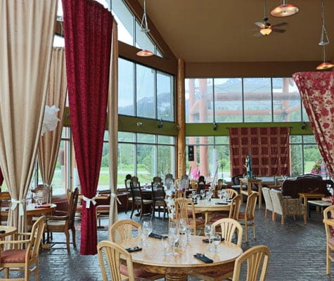 The dining area of the Tandoor & Grill, the onsite restaurant at the Stanford Fernie Resort showcases round beige wood dining table and chair sets and red, beige and red and gold print privacy curtains suspended from rods above.