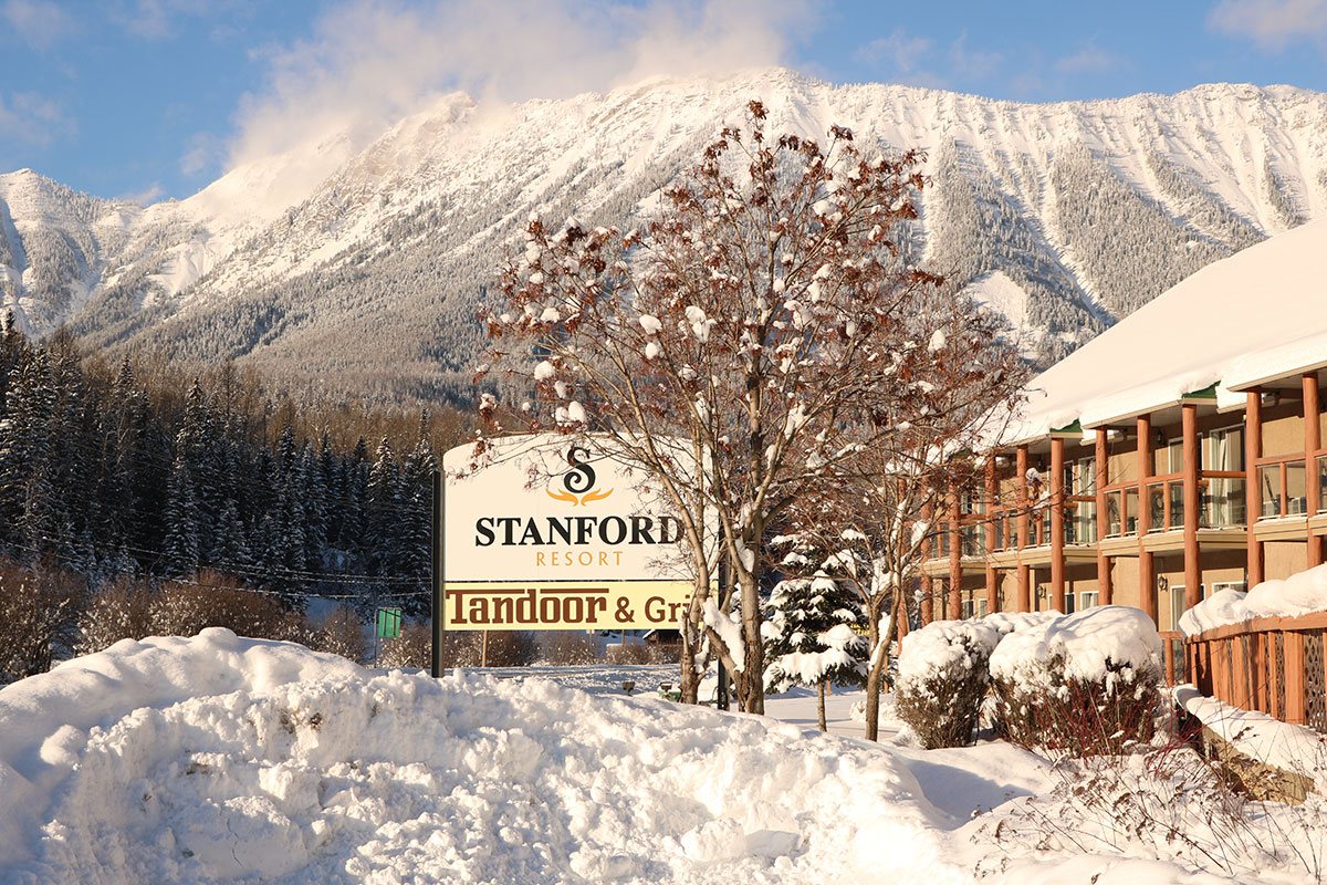 The Stanford Fernie Resort and Tandoor & Grill sign stands amongst snow covered trees, snow banks and the snow-capped Rocky Mountains in the background.