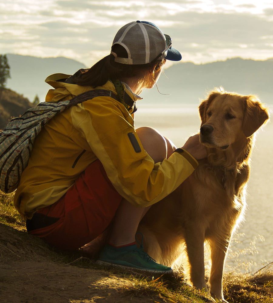 A young female wearing a baseball cap, yellow jacket and red shorts sits by her pet golden retriever looking out over the Rocky Mountains on a sunny day in Fernie, BC.