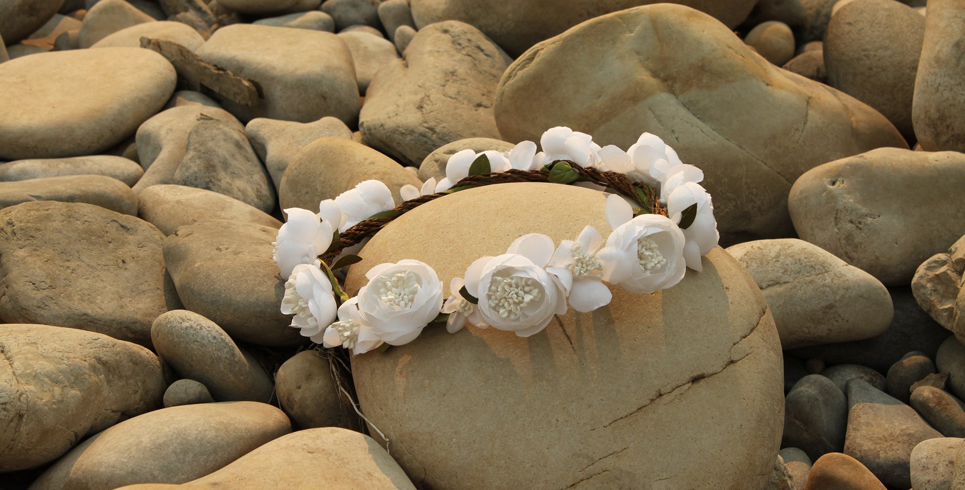 Pure white roses tied together in a bridal headband are placed amongst smooth cobbles and stones.