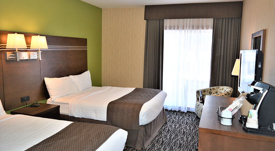 The Deluxe Room features a queen sized bed covered in a flower print quilt, two solid brown wood bedside tables, and large floor to ceiling glass patio door leading outside to a balcony.