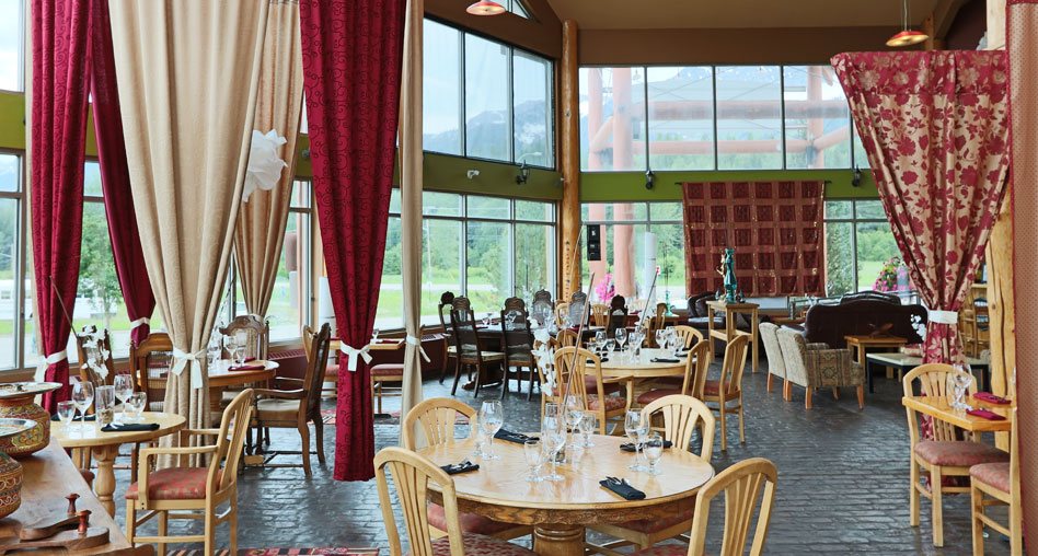 The dining space of the Tandoor & Grill Restaurant, onsite at the Stanford Fernie Resort, features floor to ceiling windows and round natural beige wood dining table sets with high-backed wood chairs to match.