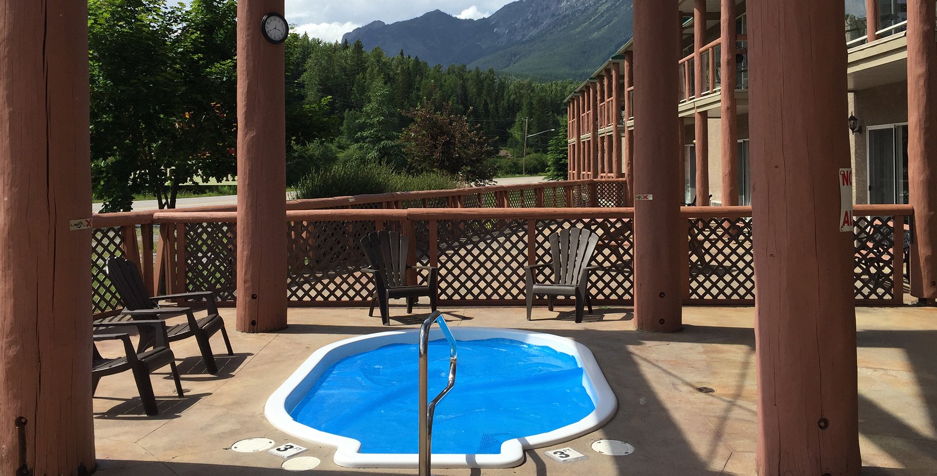 Four log posts surround the outdoor whirlpool area, at the Stanford Fernie Resort, featuring a round tub filled with crystal blue water, with four chocolate brown colored patio chairs placed around it.