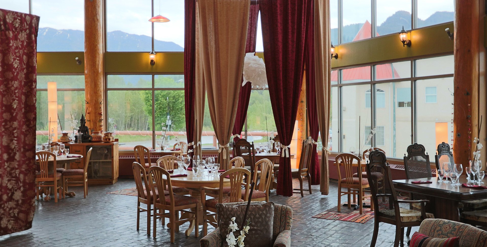 The dining room of the Tandoor & Grill Restaurant, located on the Stanford Fernie property showcases elegantly set tables placed before floor to ceiling windows reflecting scenic views of the Rocky Mountains with red and beige privacy curtains suspended from above.