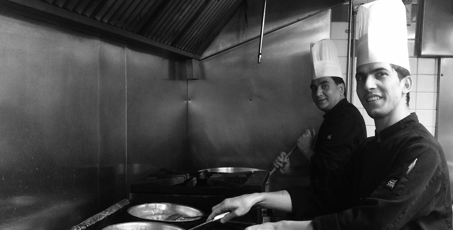 A black and white photo of two chefs wearing tall chef's hats at work over gas ranges and skillets in the kitchen at the Tandoor & Grill, the onsite restaurant at the Stanford Fernie Resort.