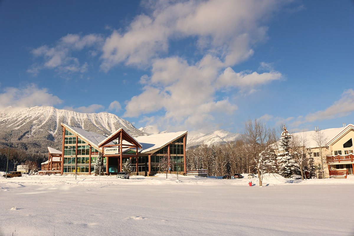 Exterior view of the Stanford Fernie Resort in the winter, with a triangular shaped portico and multi gabled roof structure of the hotel in the foreground and the snow covered Rocky Mountains in the background.