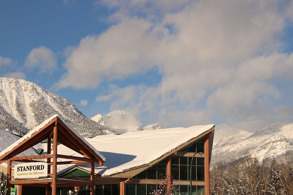 A view of the snow covered triangular roofs of the portico and the Stanford Fernie Resort pointing northward with billowing white clouds above and the Rocky Mountains blanketed in snow in the distance.