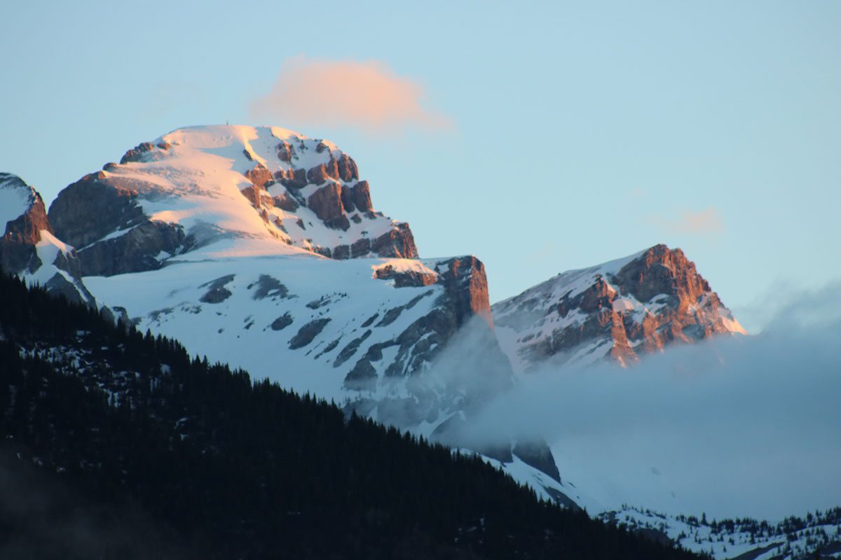 A view of the summit of the snow capped Rocky Mountains in winter with swirling white clouds and a swath of pine trees in the foreground in Fernie, British Columbia.