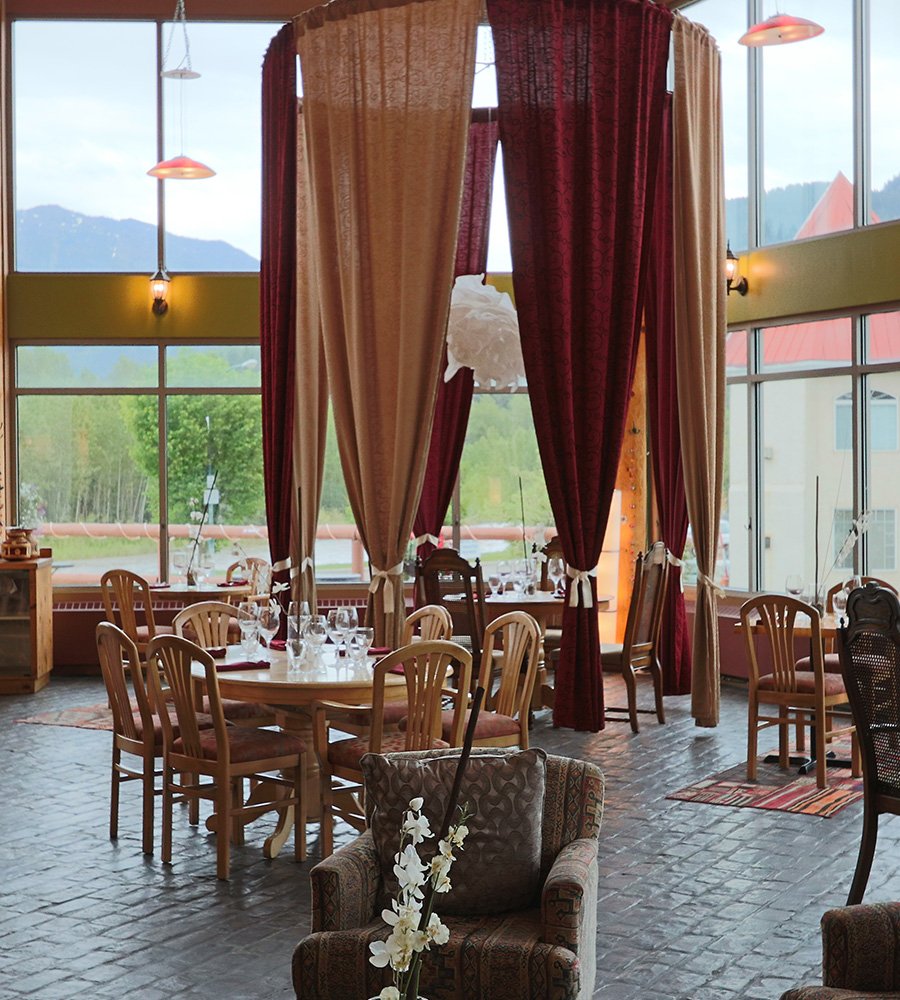 In the dining room of the Tandoor & Grill Restaurant, located onsite at the Stanford Fernie Resort, red and beige privacy curtains surround a table placed before floor to ceiling windows looking out toward scenic views of the Elk River banks in BC.