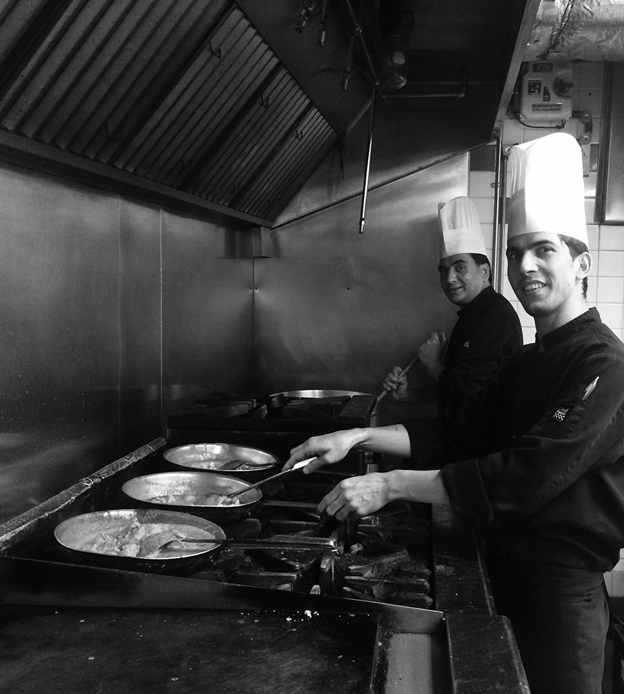 A black and white photo of two chefs preparing skillets of food over gas stove ranges at the Tandoor & Grill Restaurant, on the premises of the Stanford Fernie Resort in BC.
