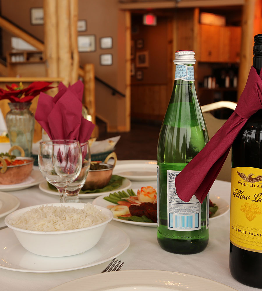 At the Tandoor & Grill Restaurant, onsite at the Stanford Fernie Resort, a table is set with bottles of mineral water and wine, wine glasses, a bowl of white rice alongside a plate of chicken tikka with garnishes.