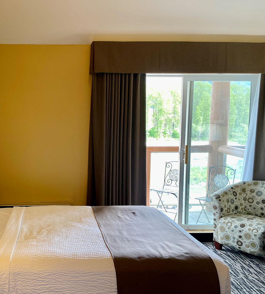 A floor lamp is lit over a moss green armchair with a coffee table bearing a plate of green grapes and cheddar cheese cubes, and two glasses filled with white wine and empty wine bottle at a suite in Stanford Fernie Resort.