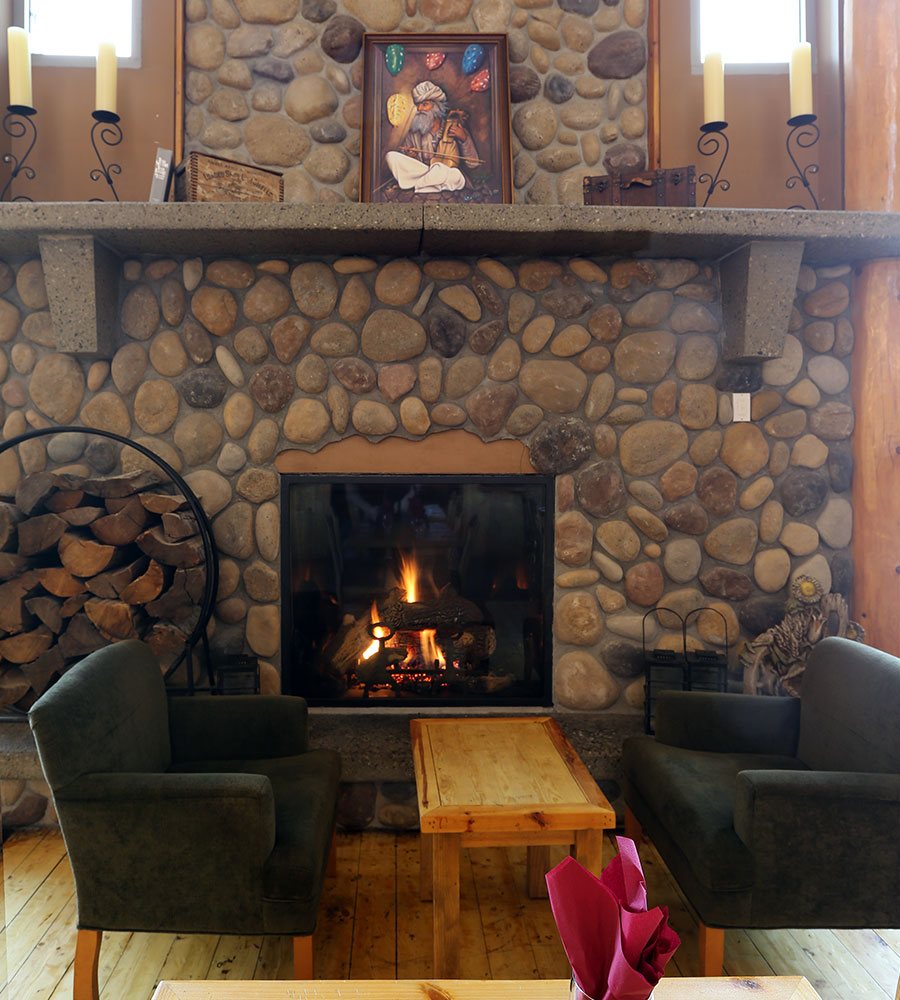 A common lounge area at Stanford Fernie Resort features two charcoal grey arm chairs placed between a rustic coffee table in front of a wood burning cobblestone fireplace.