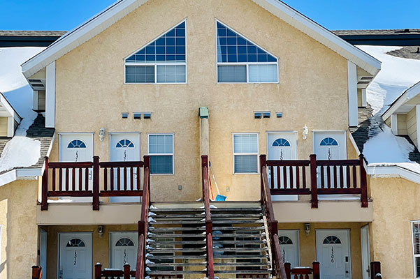 Snow covered triangular roof structures of multi storey condo units at the Stanford Fernie Resort with a steel rail staircase leading to front door entrances sheltered behind a wood guardrail.