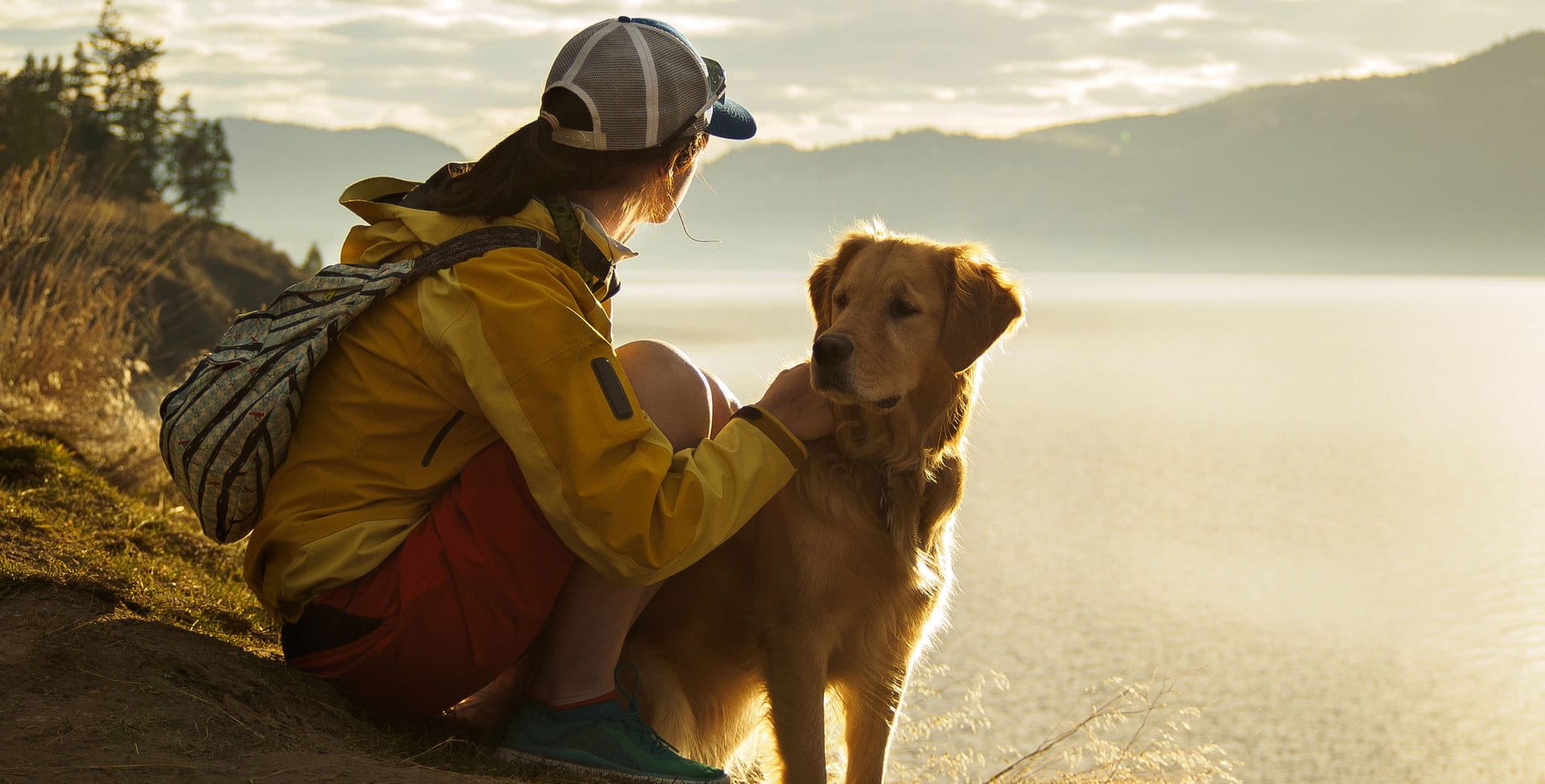 A young woman wearing a baseball cap, yellow jacket and red shorts looks out over a lake and mountains to the distance with her pet golden retriever by her side.