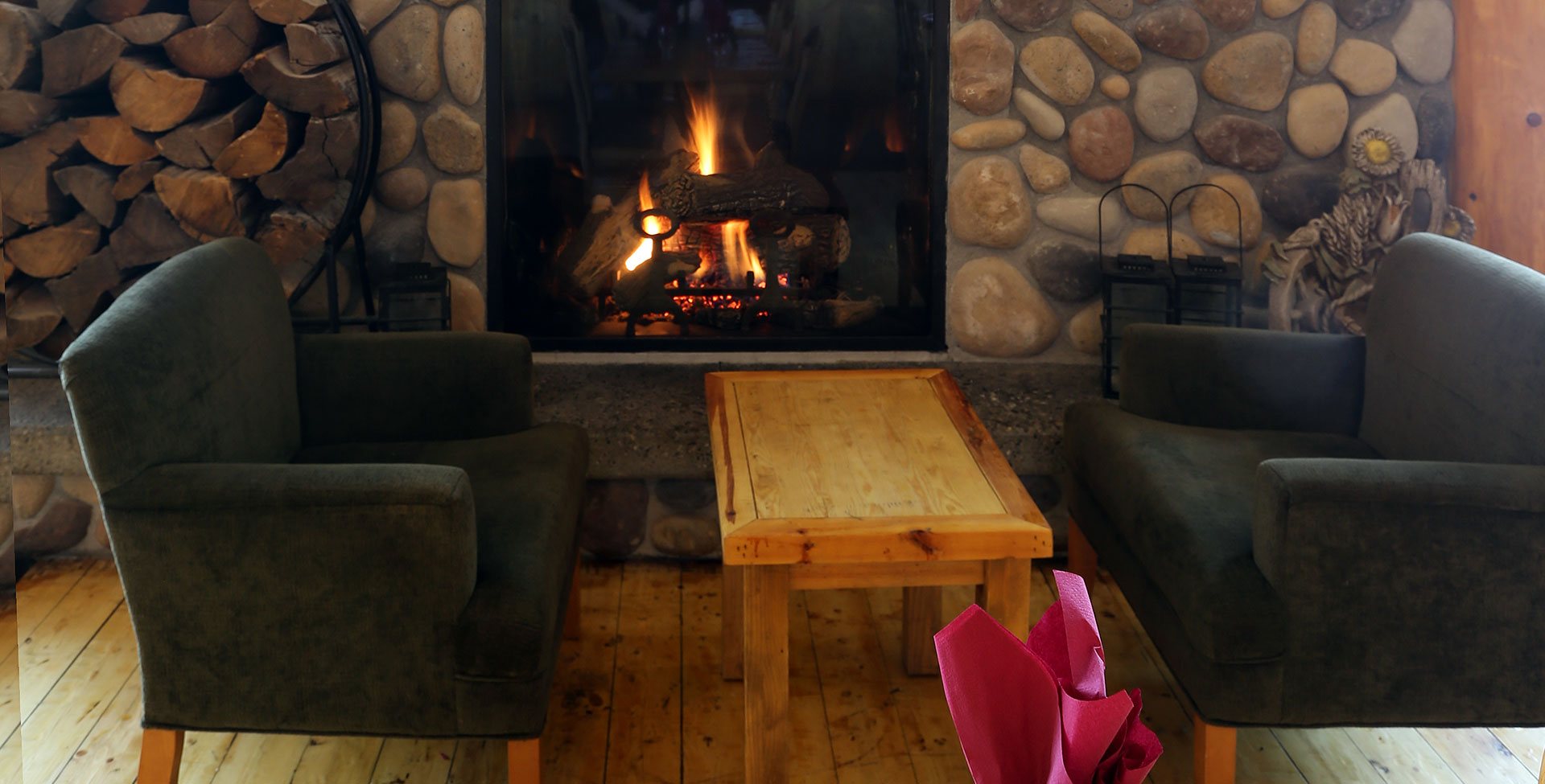 At the Stanford Fernie Resort, two charcoal colored arm chairs sit between a rustic square coffee table that are part of the lounging area with a stone woodburning fireplace.