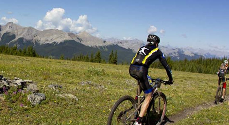 Two male mountain bikers on a bike trail ride past green pastures and rocks against a view of the snow capped Rocky Mountains in the distance in Fernie, BC.